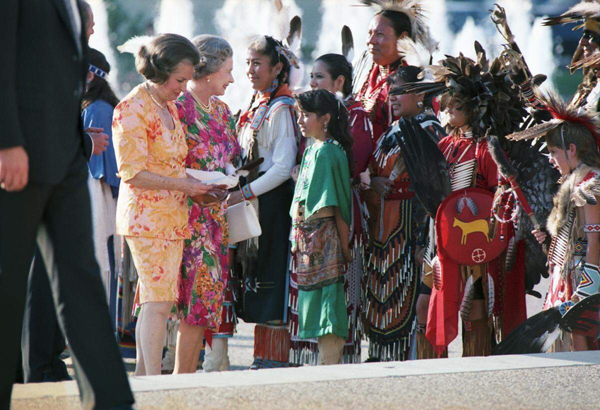 Queen Elizabeth smiles at a group of people dressed in traditional Native American attire, representing the Texas American Heritage Society, after accepting a gift from the group outside of the Hall of State in downtown Dallas.