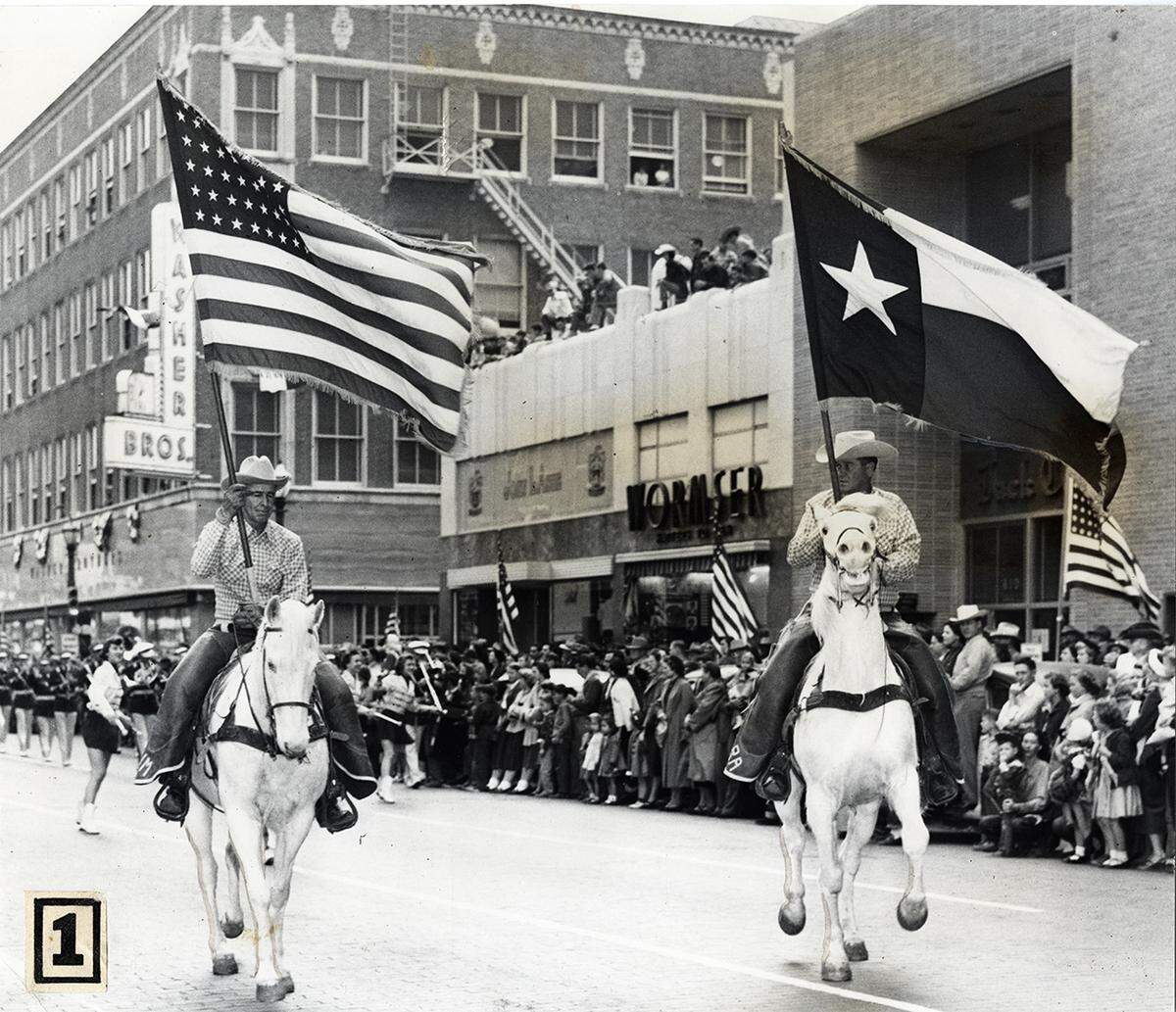 February 1953: Two men on white horses in Stock Show parade carrying U.S. and Texas flags in downtown Fort Worth.