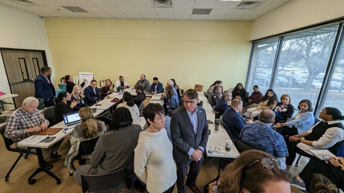 A wide angle shot of a classroom with a man and a woman in the foreground in surrounded by about 20 people working at tables. 
