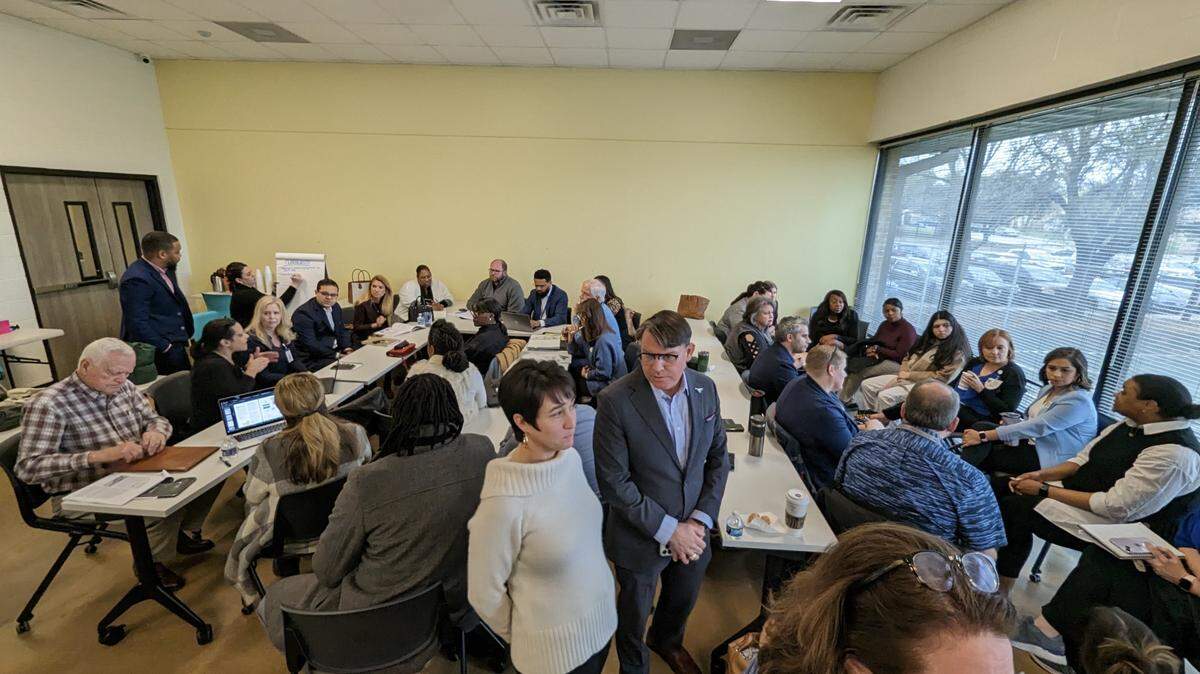 A wide angle shot of a classroom with a man and a woman in the foreground in surrounded by about 20 people working at tables. 