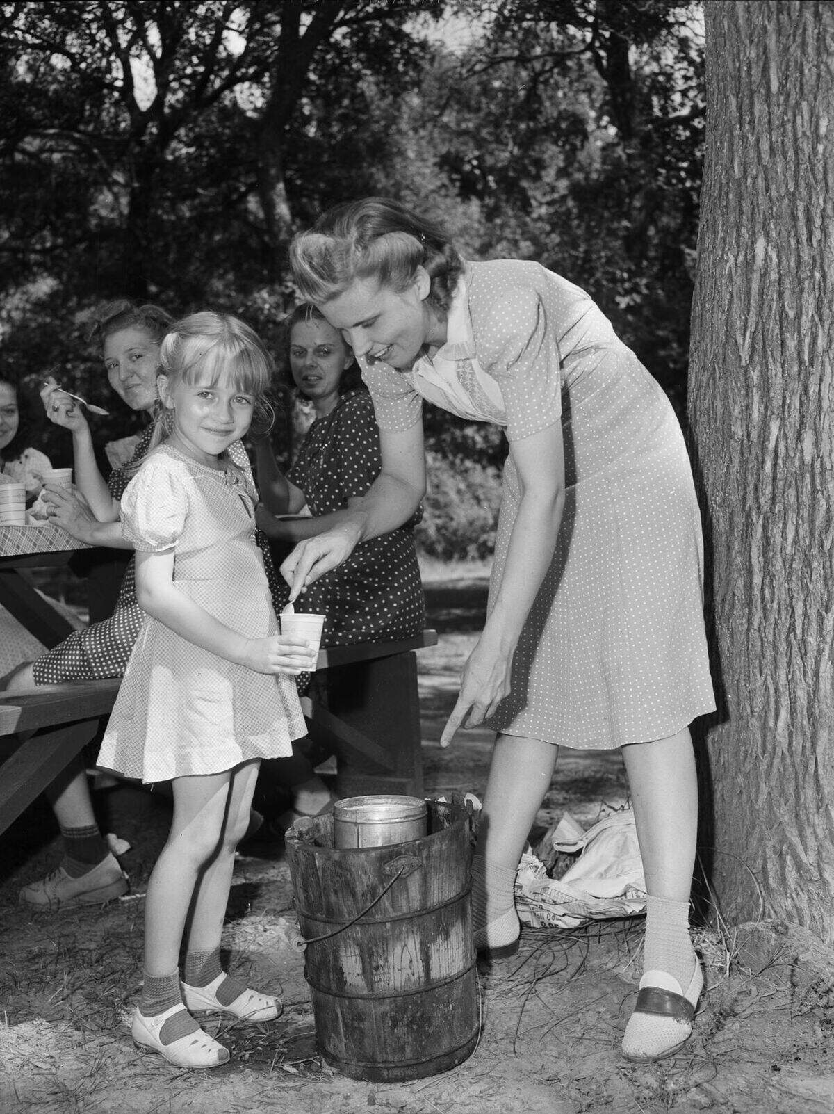 Carol is wearing a checkered dress. Miss Reed is wearing a polka-dot dress. Several girls are enjoying the ice cream at a picnic table in the background.