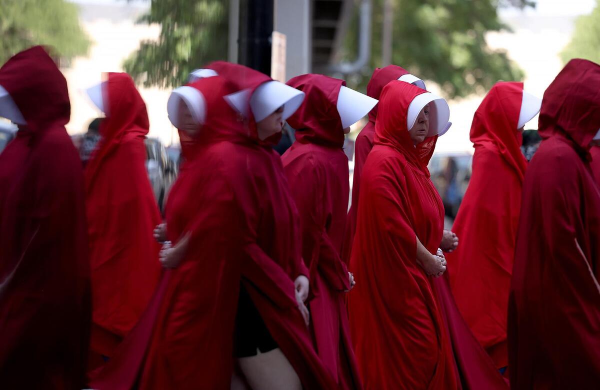 Protesters dressed in Handmaids outfits are reflected in a glass window while marching in downtown Fort Worth in the March of Dissent hosted by Indivisible 12 on June 14, 2025.