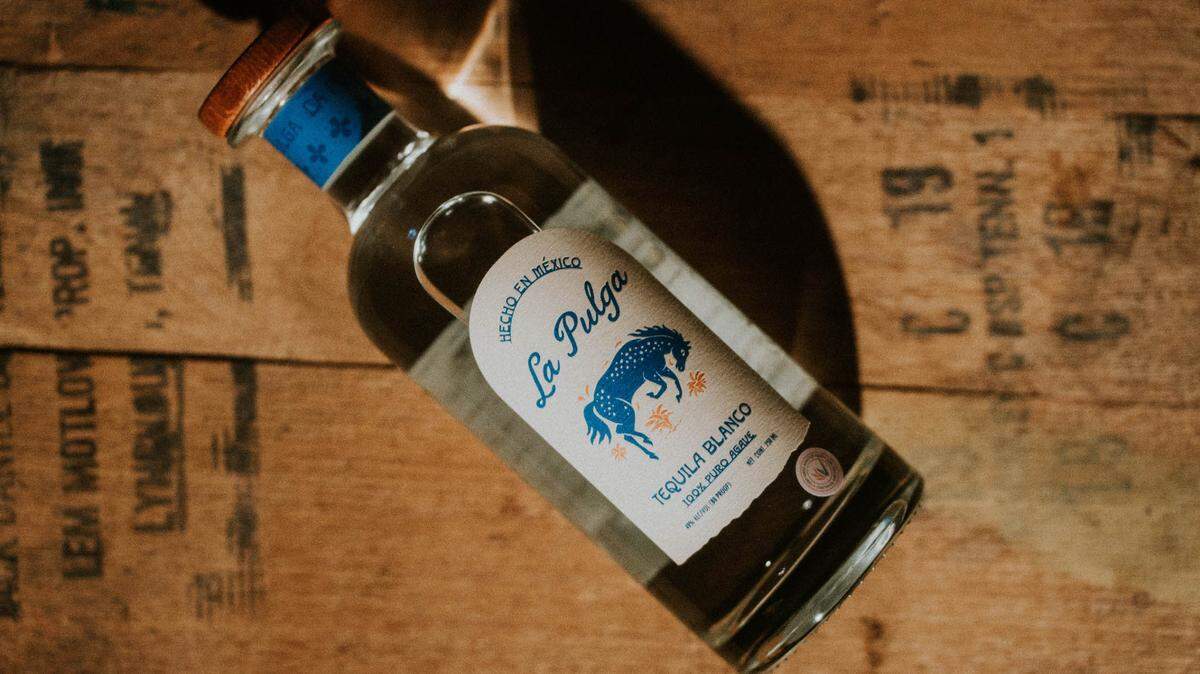 A glass bottle lying down on wooden with the La Pulga label facing up. The label reads, "Hecho en Mexico, La Pulga, Tequila Blanco" and has an image of a bucking bronco.