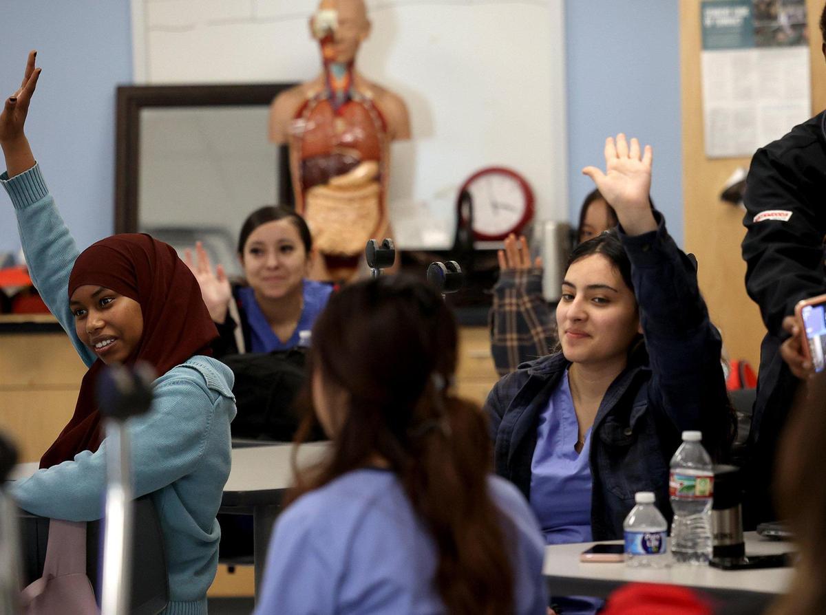 High school students in medical scrubs raise their hands, anatomy mannequin in the background.