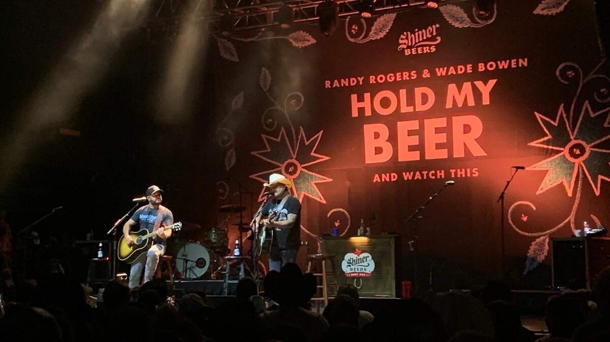 Two male lead singers perform on stage with guitars in front of a backdrop that reads quote Randy Rogers and Wade Bowen: Hold My Beer.
