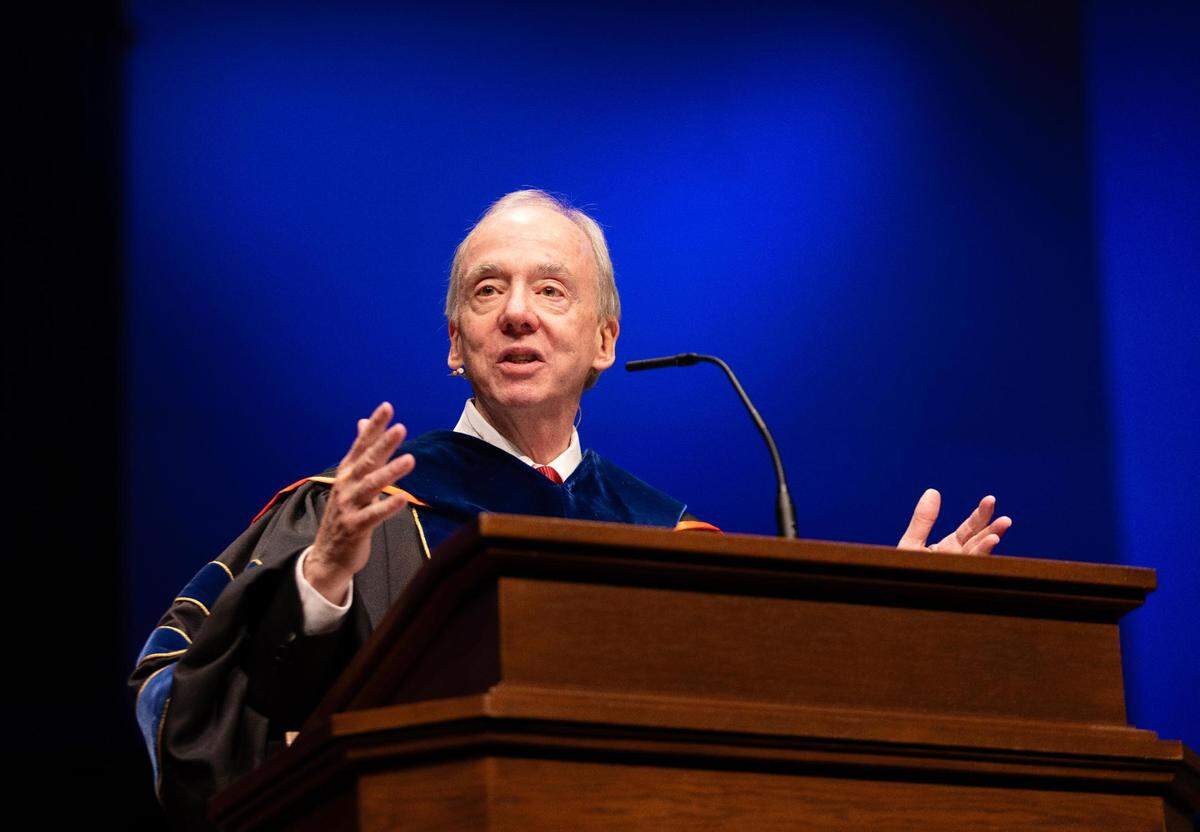 An older man in a graduation gown stands at a podium while speaking, his arms slightly raised, with a blue background.