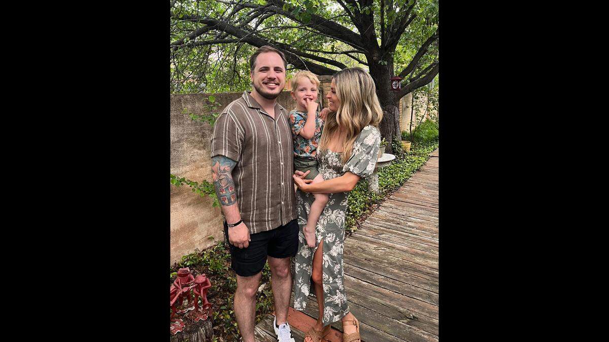 The family of three is pictured smiling in front of a tree and surrounding greenery.