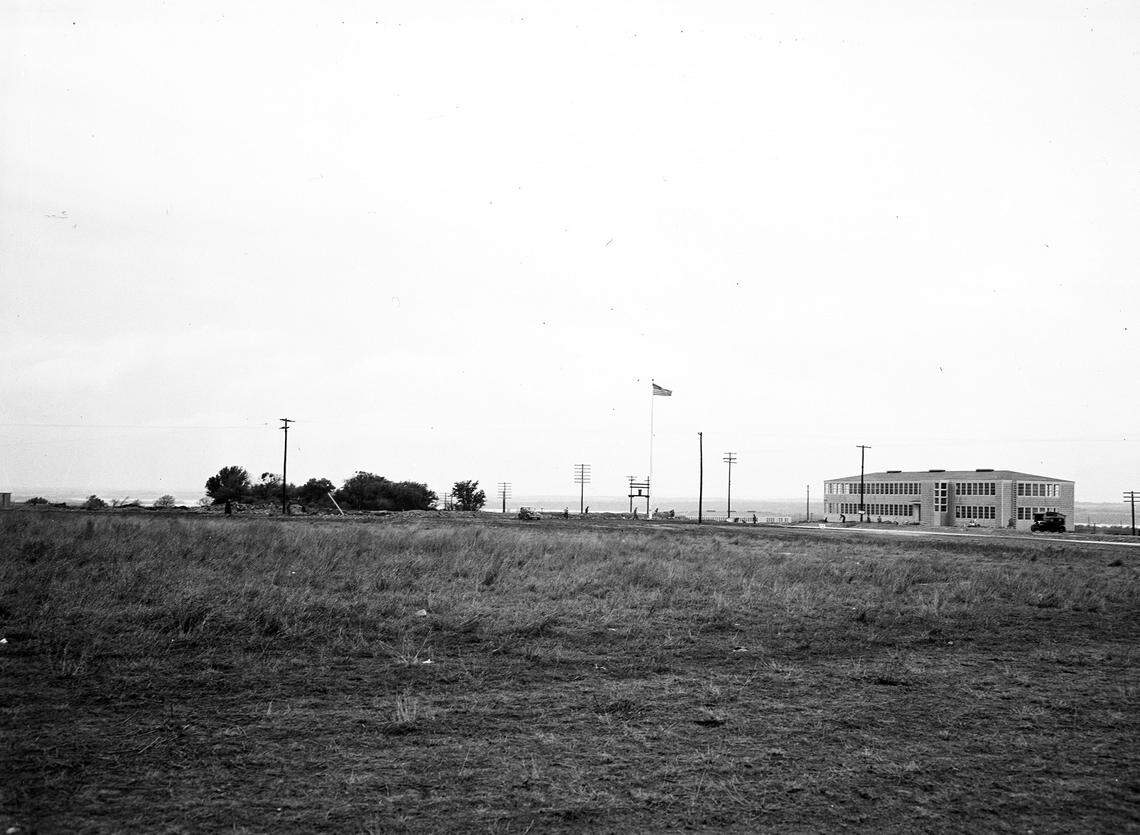 A building is standing toward the center-right of the photograph, behind it, flat Texas planes give way to the horizon. Several people seem to be making their way in and out of the building, perhaps to or from the automobile that that parked on the street just outside of the building. Electric poles/pylons are dotting the scene. A tall flagpole is standing taller than everything else, with a United States flag flying at full-staff.