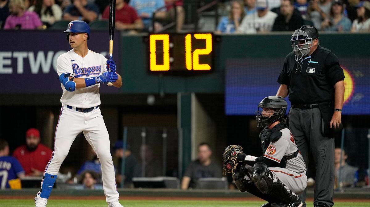 Texas Rangers’ Nathaniel Lowe, left, Baltimore Orioles catcher Adley Rutschman and umpire Hunter Wendelstedt stand ready for a pitch as the pitch clock counts down during a baseball game. Lowe is one of five Rangers with a Batter Pitch Timer Violation.