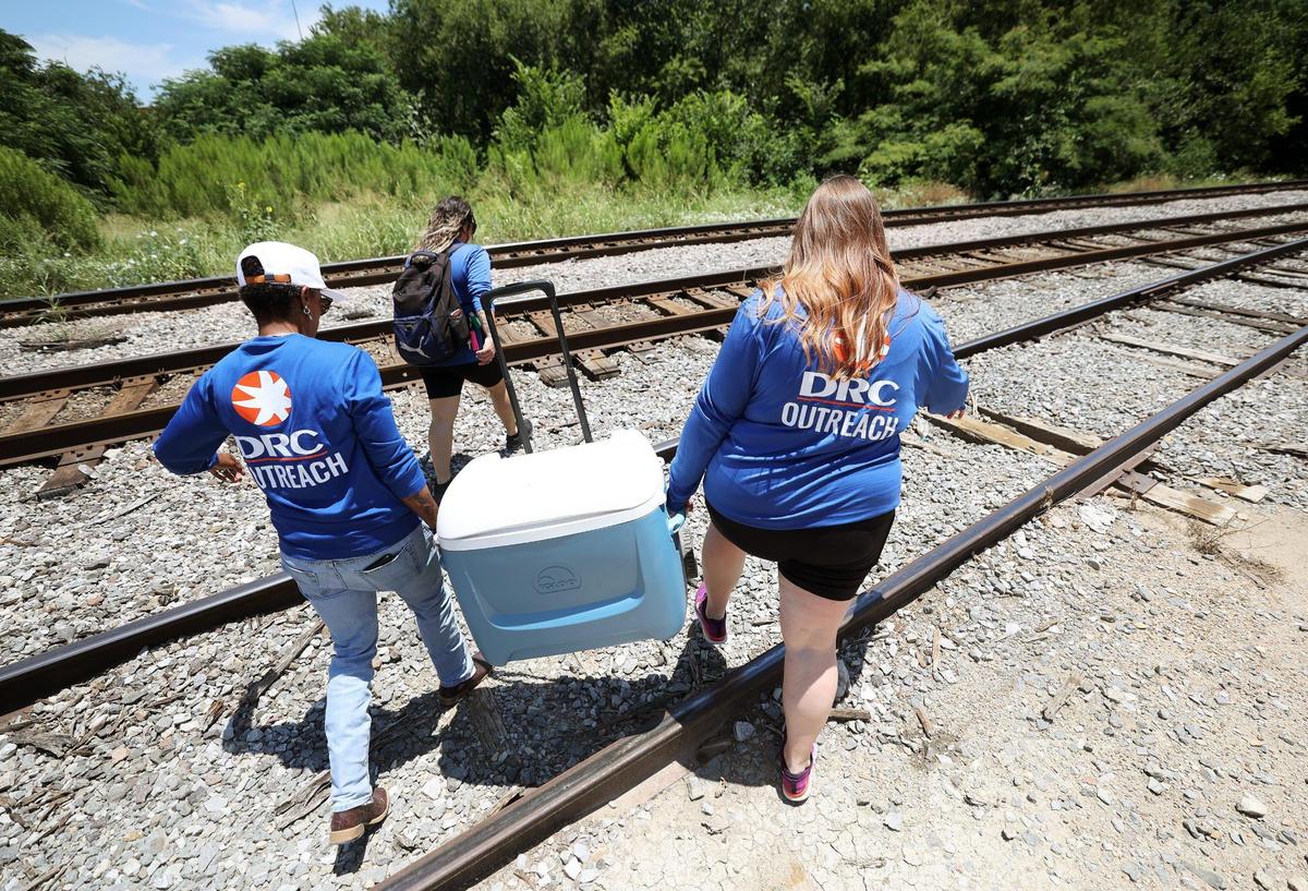 People wearing DRC Outreach shirts carry a water cooler over a railroad track towards an area covered with trees.