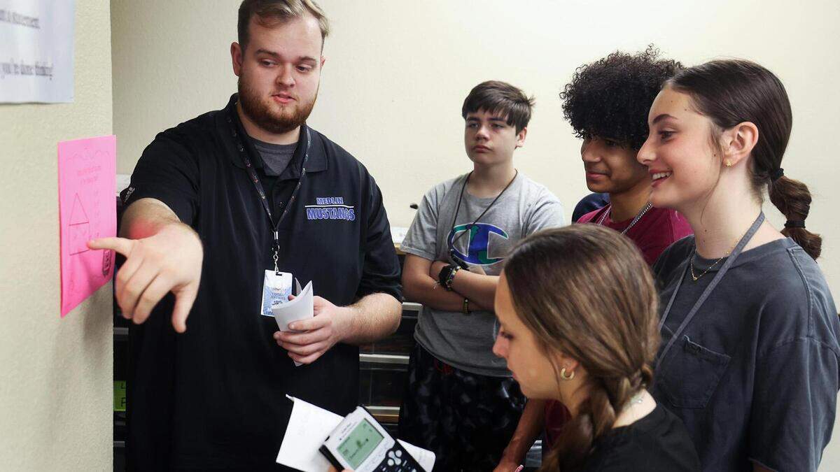 A man in a black polo shirt points to a pink piece of paper taped to a wall. The paper has a math problem on it. Four eighth graders look at the paper as the man explains.