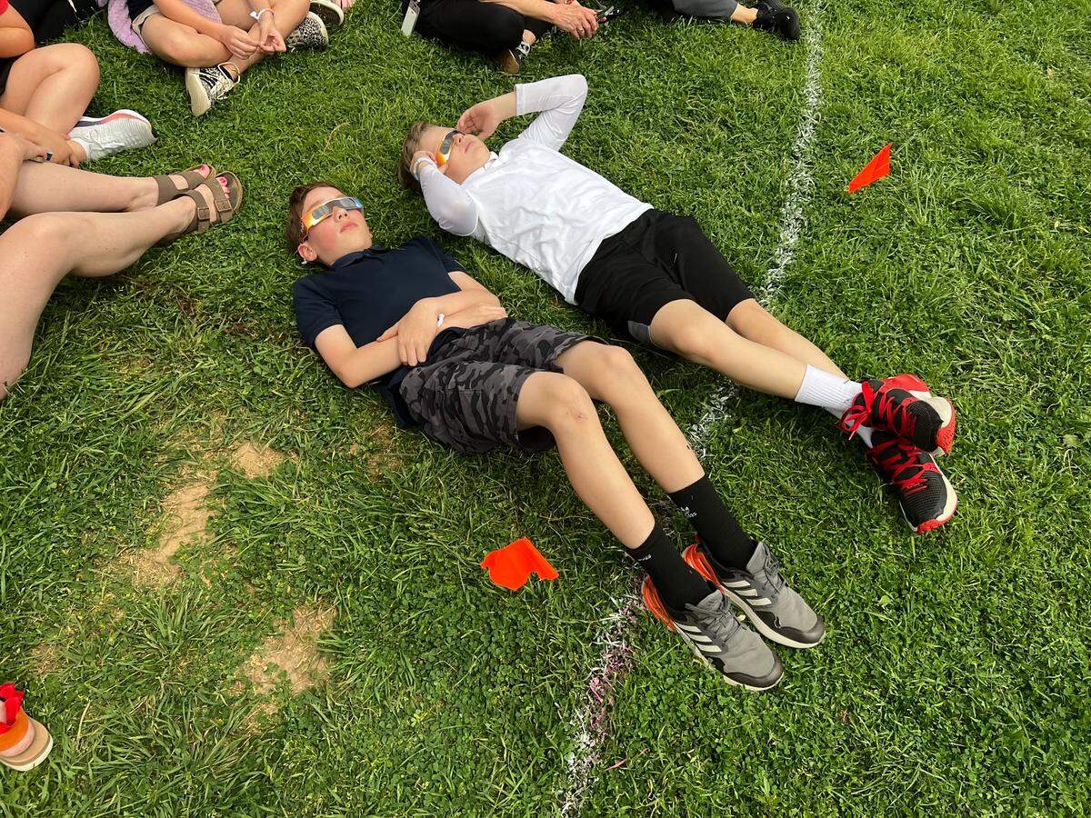 Two elementary students are lying on a grassy field while staring at the sky and sporting their protective eyeglasses. 