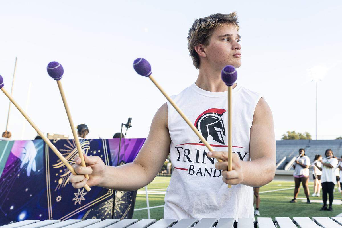 A young man in a Trinity band t-shirt with the sleeves cut holds two mallets in each hand while playing the metal slots of the vibraphone. 