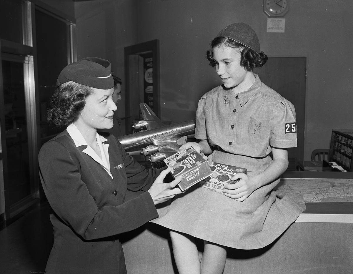 March 18, 1954: Miss Mary McAlpin, American Airlines stewardess, examines a box of the Girl Scout cookies purchased for all flights of the lines out of Carter and Love Field. Showing her the cookies is Brownie Mary Frances Archer.