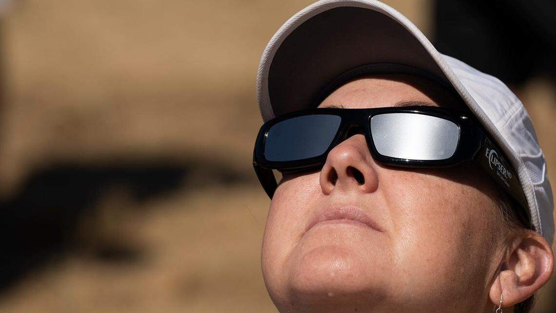 A woman looks up into the sky wearing eclipse glasses with dark lenses. Her face is looking up into the sun, and is covered in sunlight. 