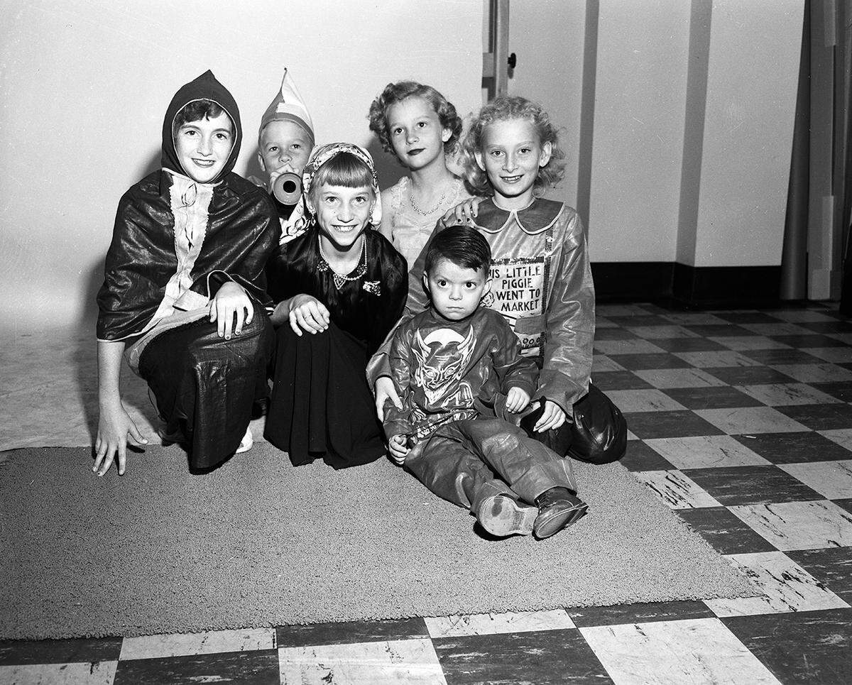 Six young children wearing costumes are huddled together and smiling at the camera on a run in what looks to be a school classroom.
