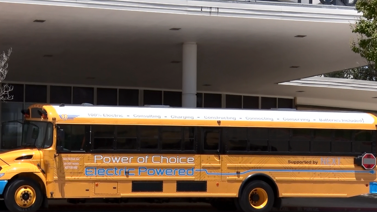 An electric school bus, which looks almost identical to a regular school bus, is pictured. It's mostly yellow but has the words "electric powered" written in blue on the side. The bus' front bumper and back bumper are also the same color of blue. 