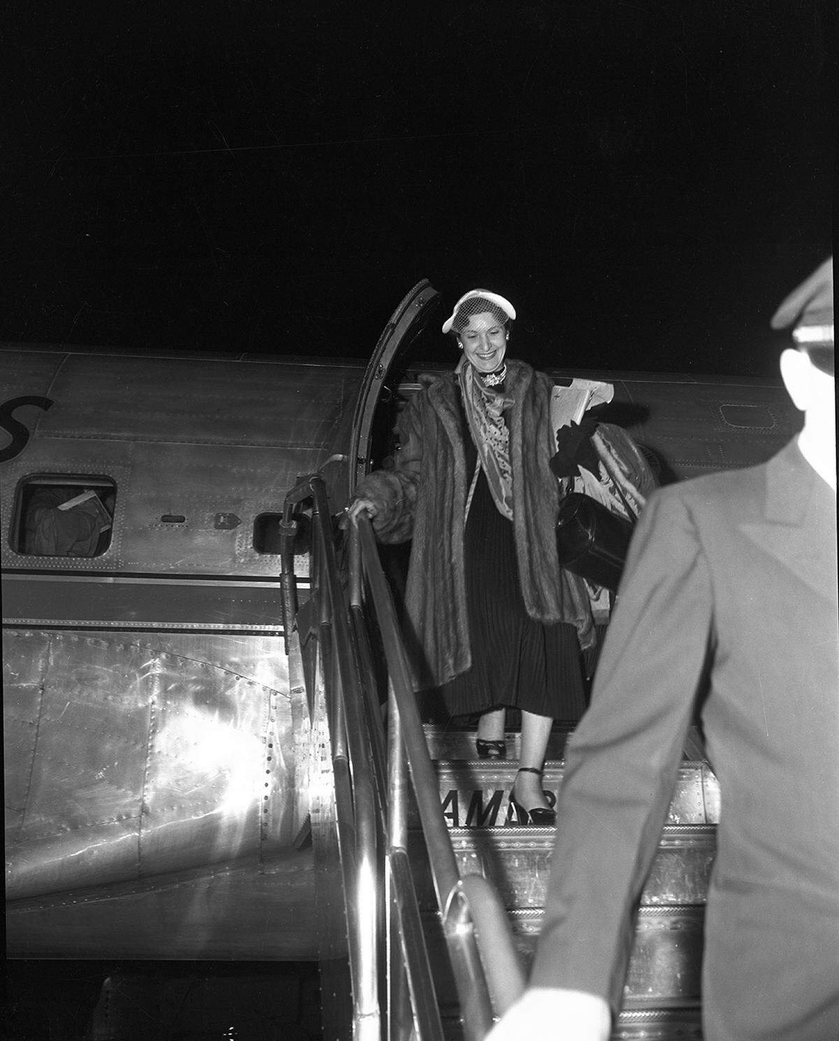 The woman is wearing a dark dress and fur coat. She is holding a large purse and smiling as she steps down the stairs.