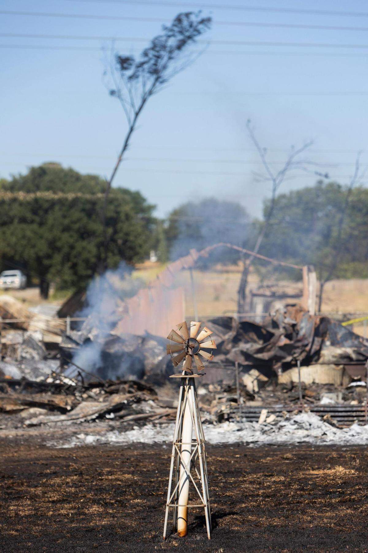 A charred windmill stands behind a small patch of burning flames after a home fire in Rendon, Texas, on Wednesday, July 27, 2022.