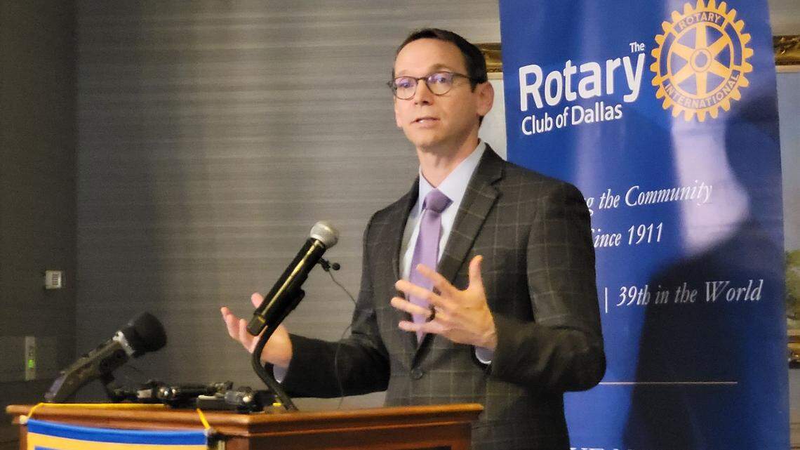 A bespectacled man gestures with both hands while standing behind a podium and speaking. Behind him is a blue banner reading "Rotary Club of Dallas."