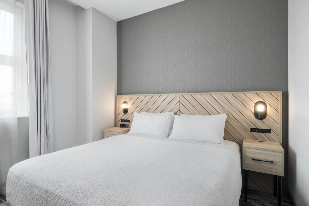 A room inside the Sandman Signature Hotel features a King size bed with a white comforter blanket and pillows with a light brown wooden headboard against grey walls.