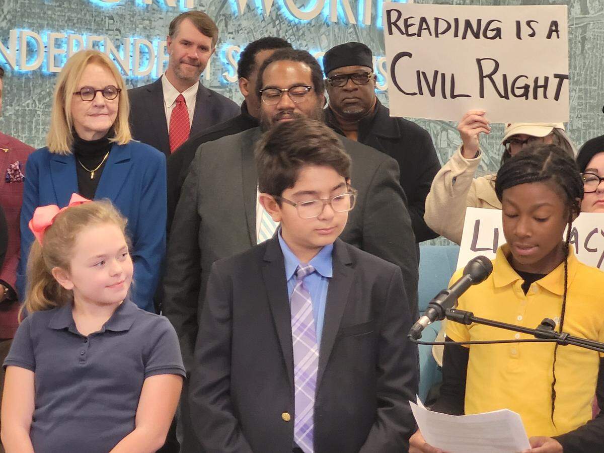 A girl in a yellow shirt reads into a microphone from a piece of paper. A boy and a girl look on. A group of adults stands behind them, one holding a sign that reads "Reading is a Civil Right."
