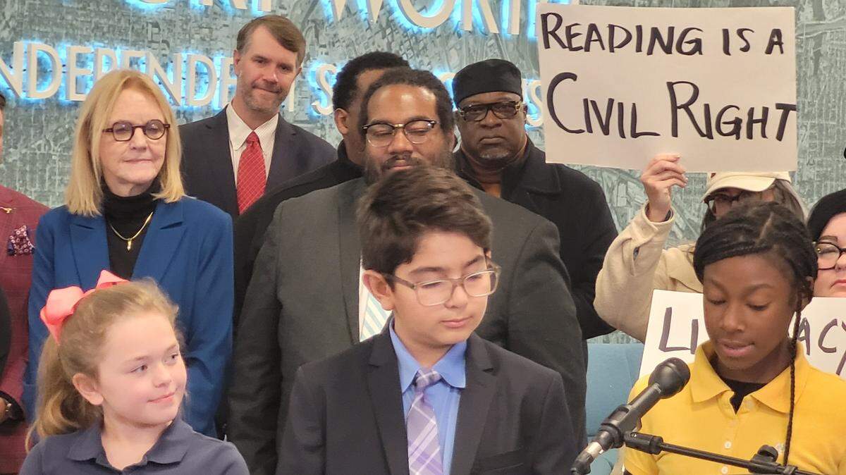 A girl in a yellow shirt reads into a microphone from a piece of paper. A boy and a girl look on. A group of adults stands behind them, one holding a sign that reads "Reading is a Civil Right."