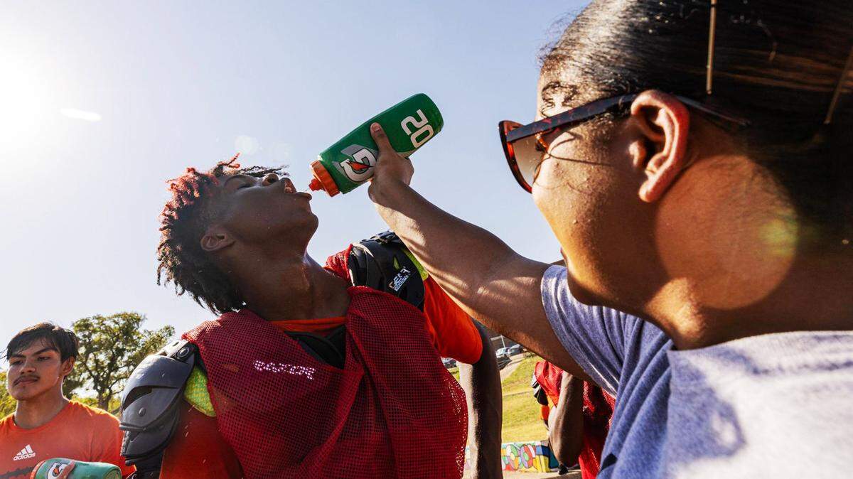 A person squirts water from a bottle into a football players mouth as the sun shines.