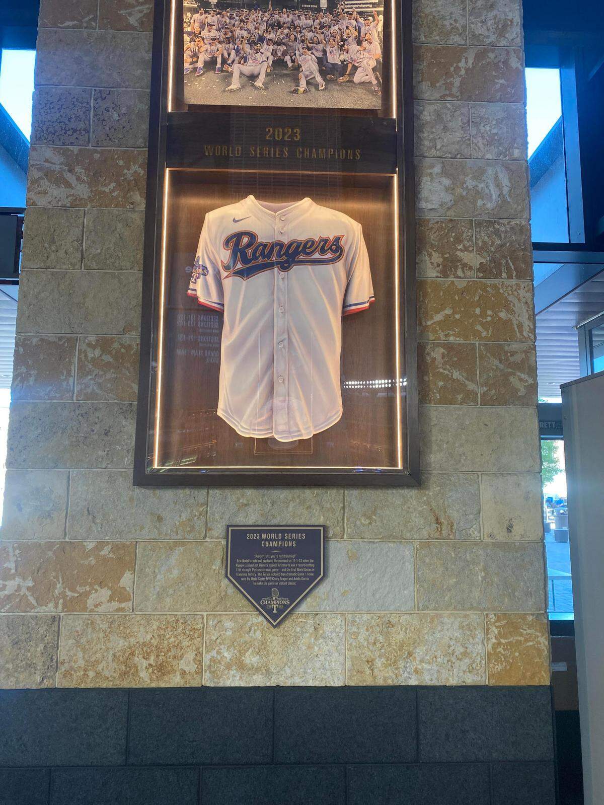 A framed Rangers World Series jersey with a picture o the team after winning the World Series on top of it and a plaque below it.