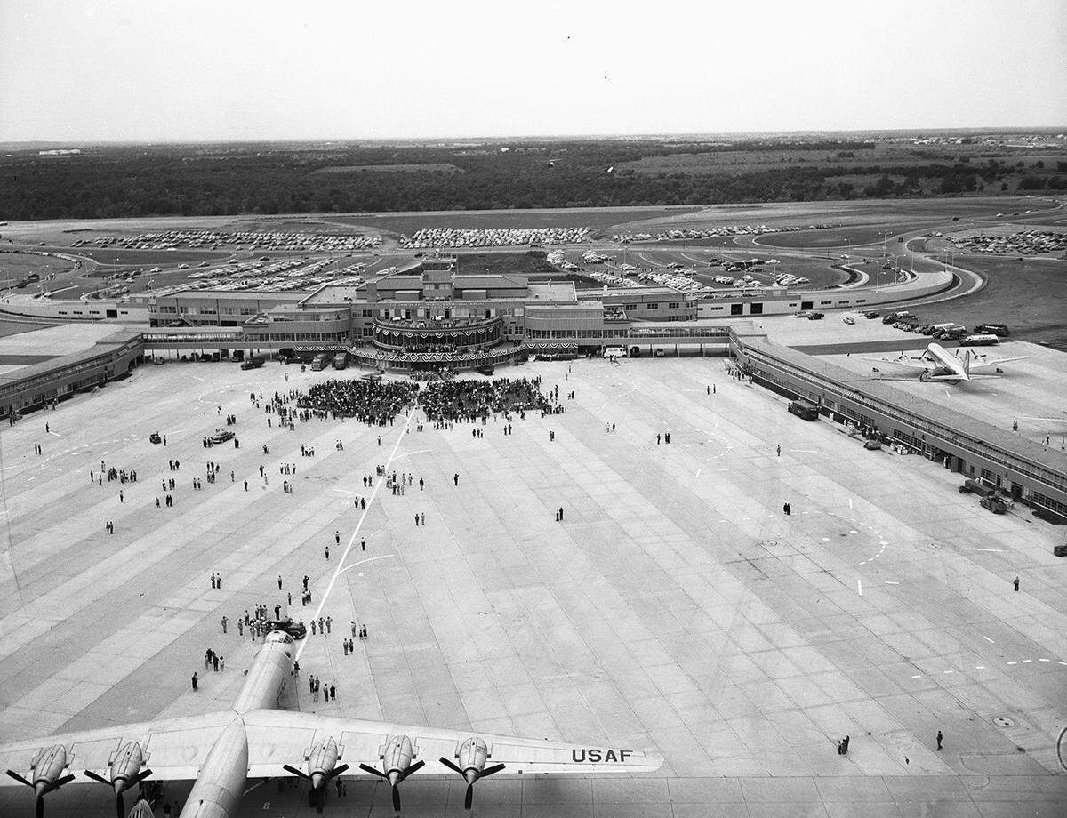 An aerial view of the terminal, runway, parked airplanes, and parked visitor cars at the event. In the foreground is a military airplane and spectators in front of the terminal.