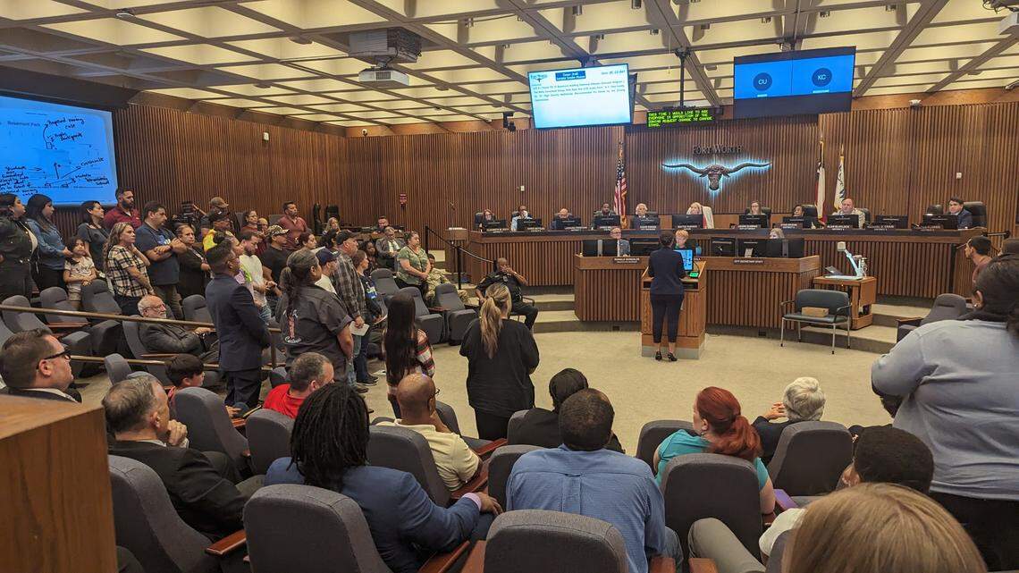 A group of people standing in a city council chamber while a person speaks at the lectern.