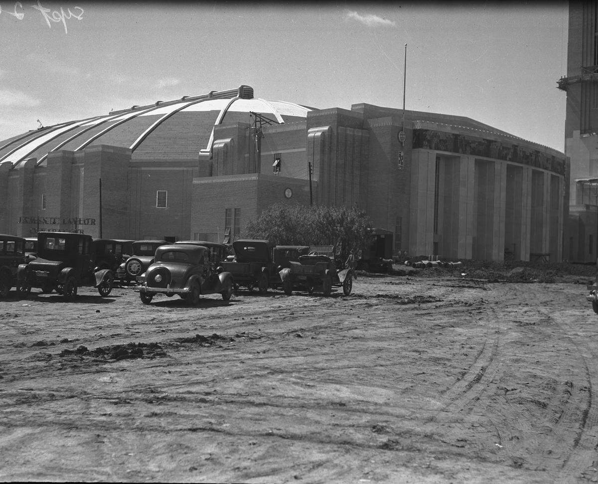 1936: Construction site of Will Rogers Coliseum complex with cars parked beside building.