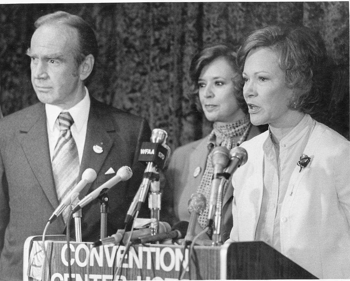 First lady Mrs. Rosalynn Carter visits Fort Worth in 1980, speaking at a convention podium to the public and press. Besides her are House Majority Leader Jim Wright of Fort Worth and his wife, Betty Wright.