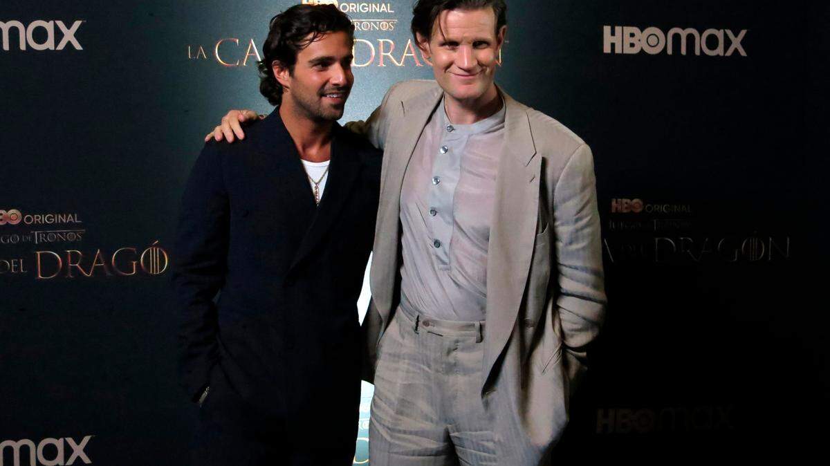 The actors pose with their arms around their shoulders, smiling in front of a banner with the show's name on it.