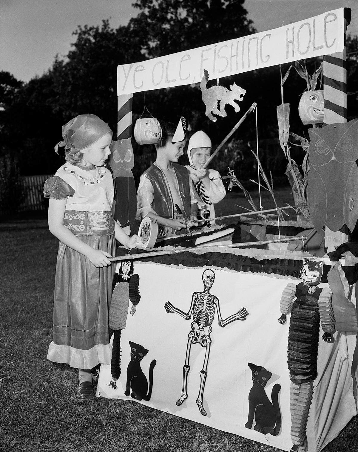 Three children wearing costumes stand around a decorated booth labeled as "Ye Ole Fishing Hole". They are smiling as they hold fishing poles over the center of the booth.