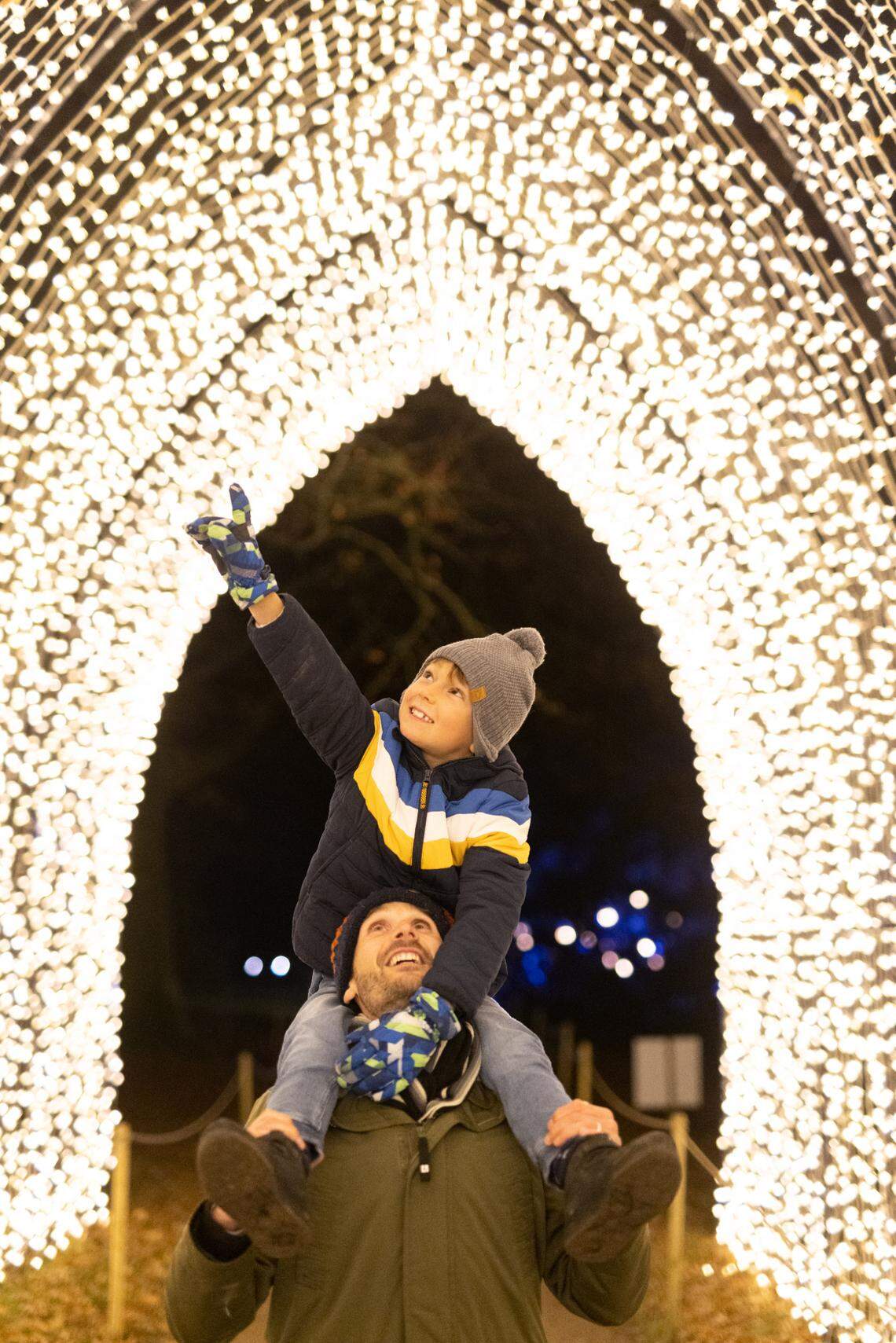 A young boy sits on top of his father's shoulders and points up at hundreds of gold lights illuminating in an arch.