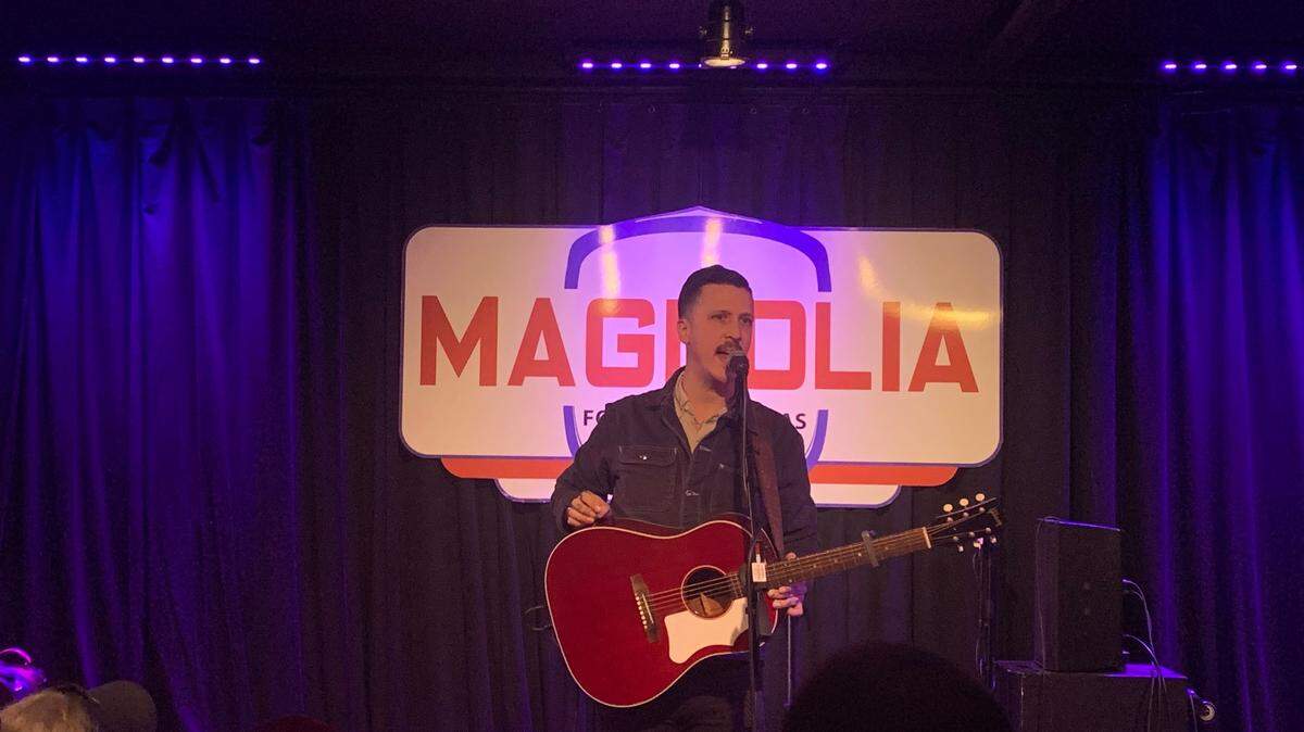A man playing a guitar stands on a stage in front of a sign that reads quote "Magnolia."