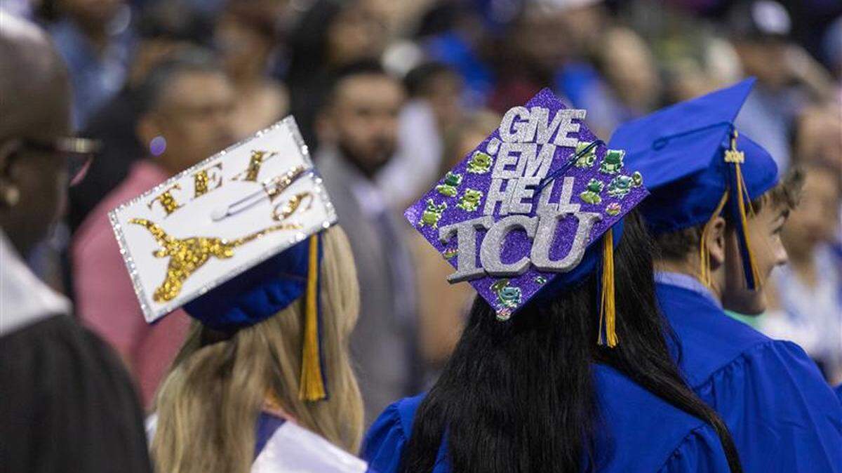 Two girls in blue graduation gowns, one wearing a mortarboard with the word "Texas" and the UT longhorn logo, the other wearing a purple mortarboard with the words "Give 'em hell, TCU."