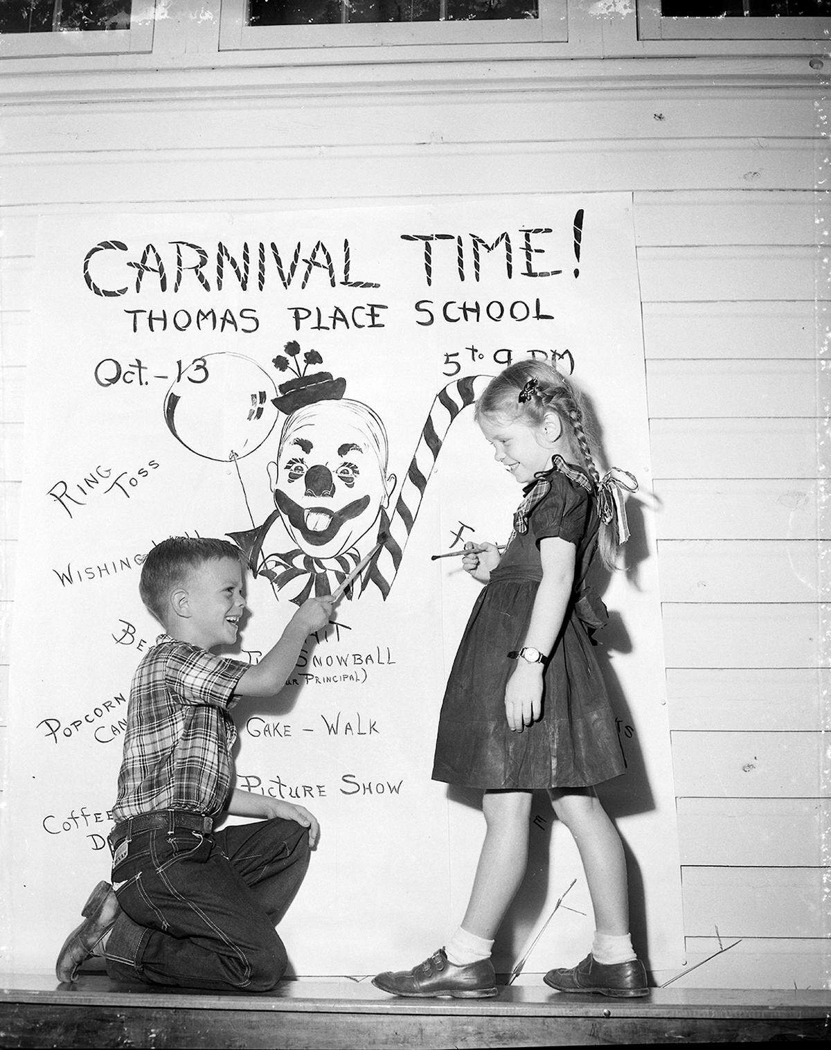 A boy in jeans and girl in a dark dress smile at each other holding paint brushes in front of a large poster that says "Carnival Time!" with a painted clown face on it.