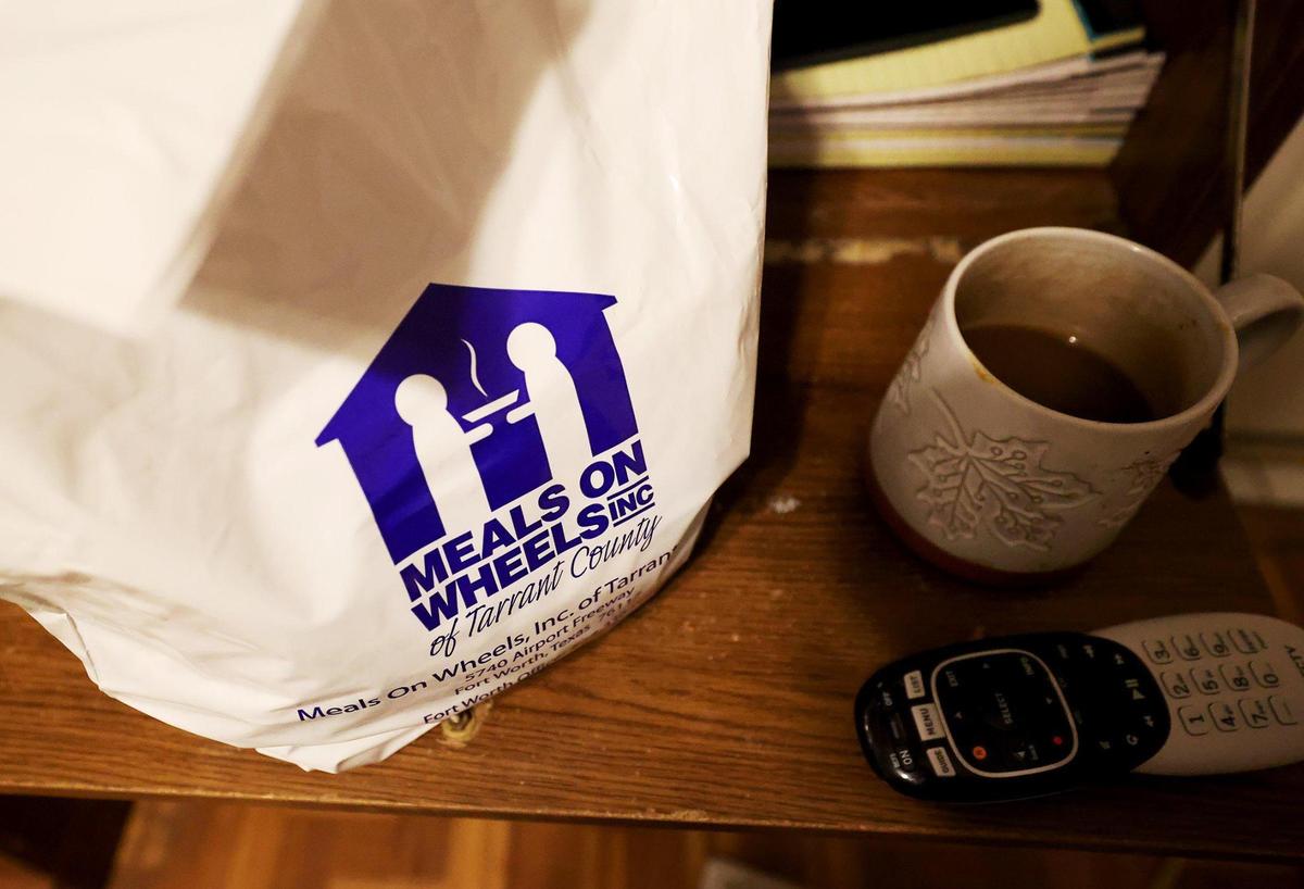 A white plastic bag with Meals on Wheels of Tarrant County sits on a wooden desk next to a cup of coffee and remote controller.