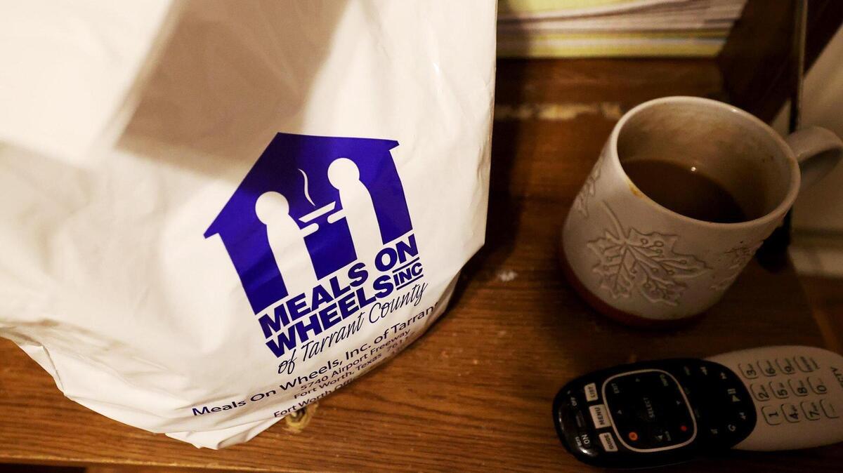 A white plastic bag with Meals on Wheels of Tarrant County sits on a wooden desk next to a cup of coffee and remote controller.