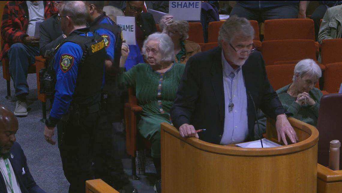 In a screenshot from the commissioners court meeting, members of the audience hold up signs reading “SHAME” in response to County Judge Tim O’Hare having a member of the public removed from the room. The man removed, Bishop Kirkland, had commented on O’Hare banning clapping by the audience.