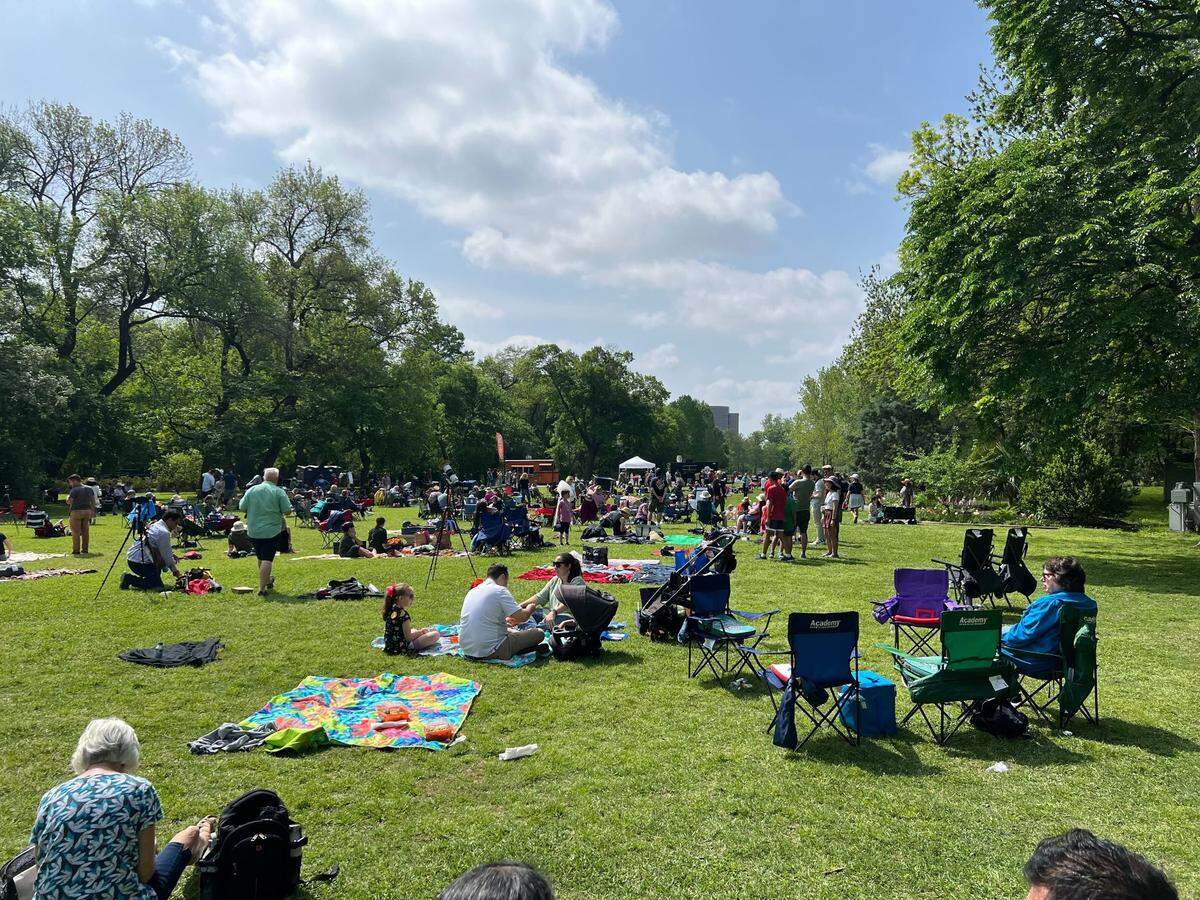 On an open lawn, dozens of people sit in chairs or on blankets as the sky appears to be sunny with some broken clouds.