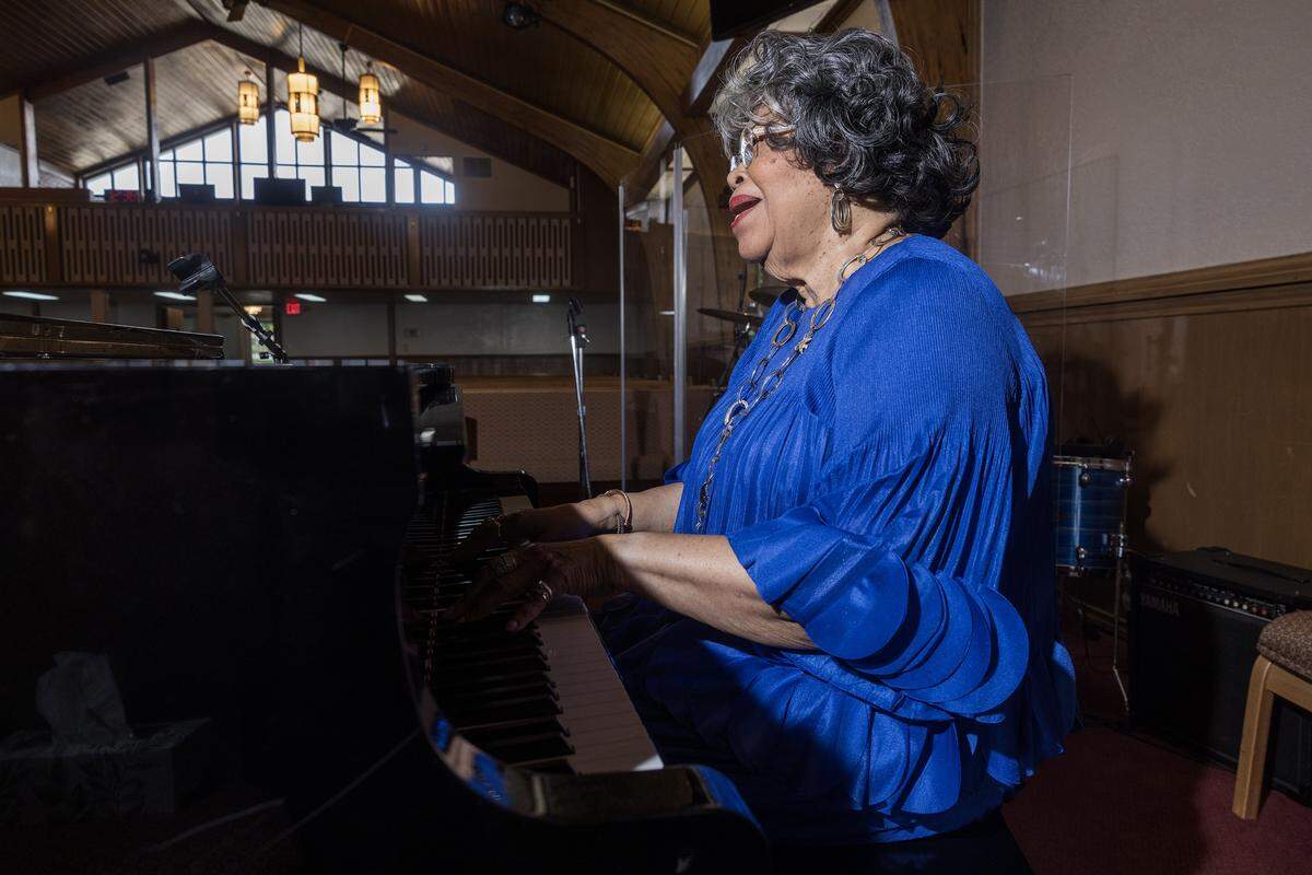 An older woman in a bright blue dress plays the piano in a darkly lit church.