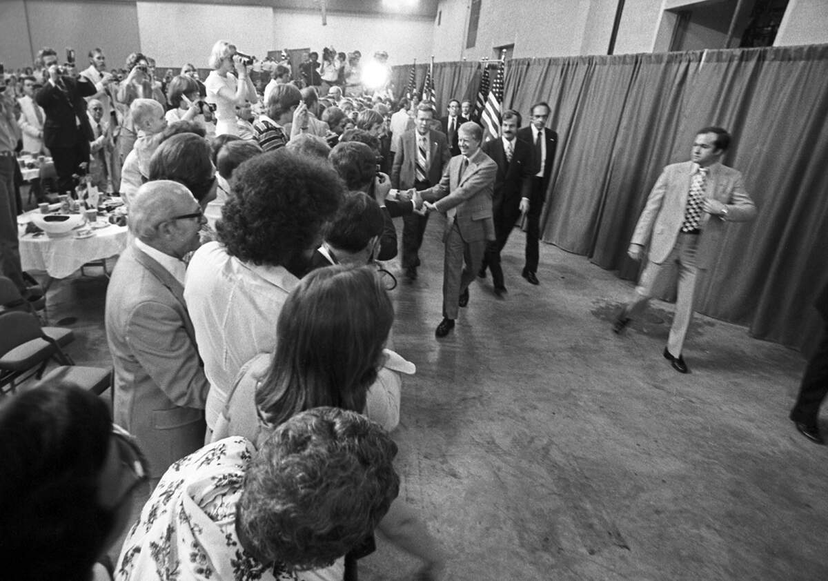 Carter walks along the edge of a crowd waving and shaking hands with people.