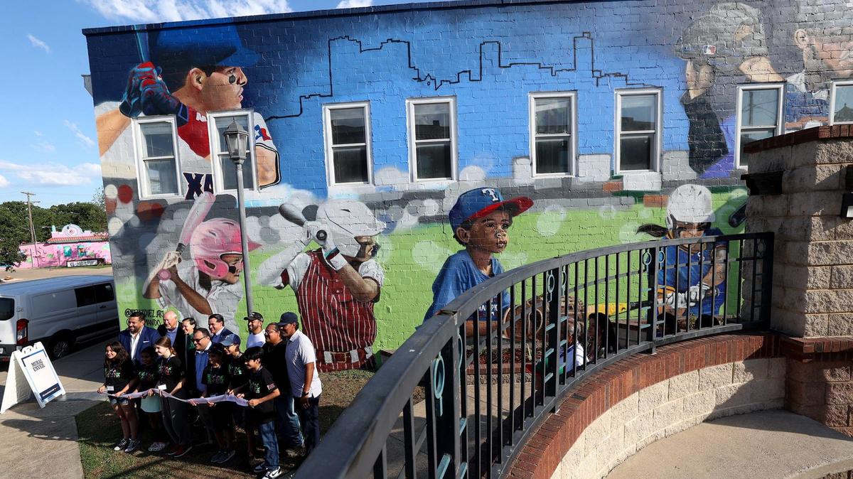 Children of the Artes de La Rosa Cultural Center, MLB officials and city representatives take photos in front of a mural celebrating MLB All-Star Week and Latino softball and baseball players A new owner plans to paint over the mural. 