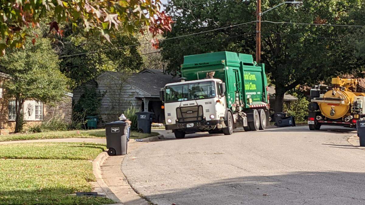A garbage truck rounds the corner in a tree lined neighborhood. 