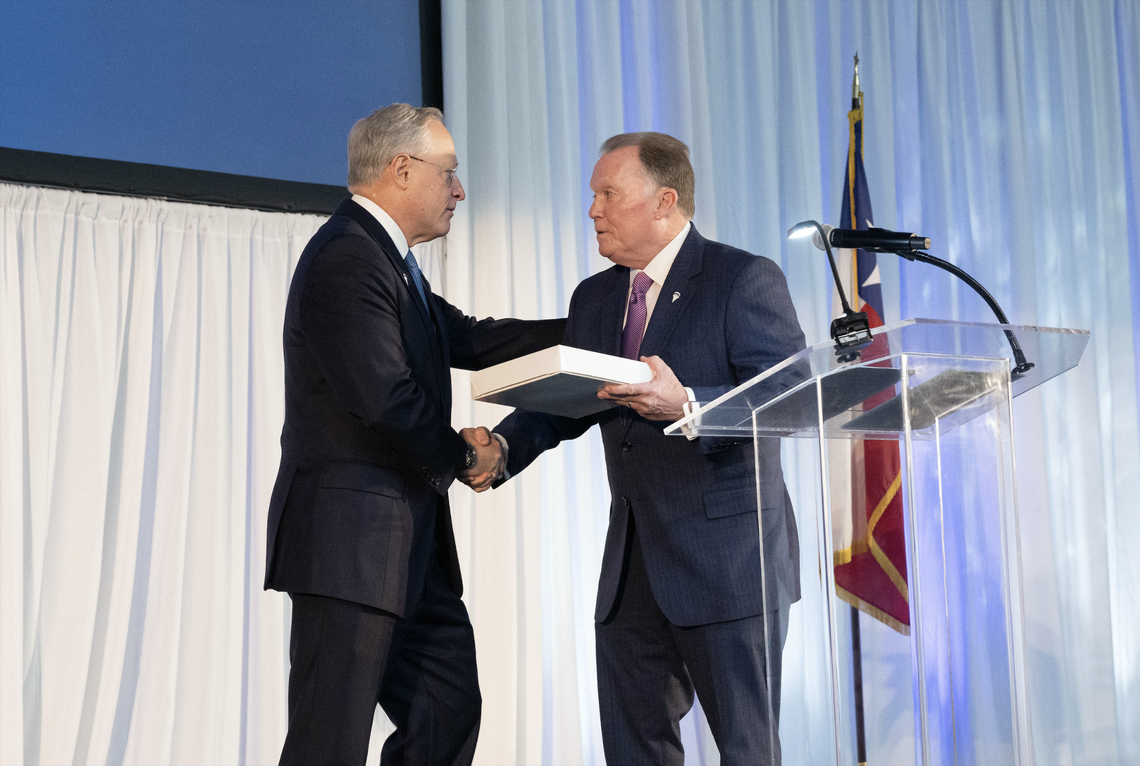 Mike Berry shakes Ross Perot Jr.'s hand while handing him a white box. Perot Jr. places his hand on Berry's shoulder as the two lock eye contact. They stand behind a clear podium in front of a white curtain, American flag and presentation screen.