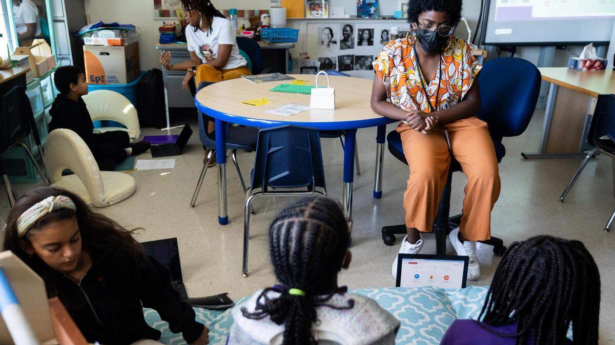 A woman in a multicolored shirt and orande pants sits at a table and reads to children who are seated on the floor.