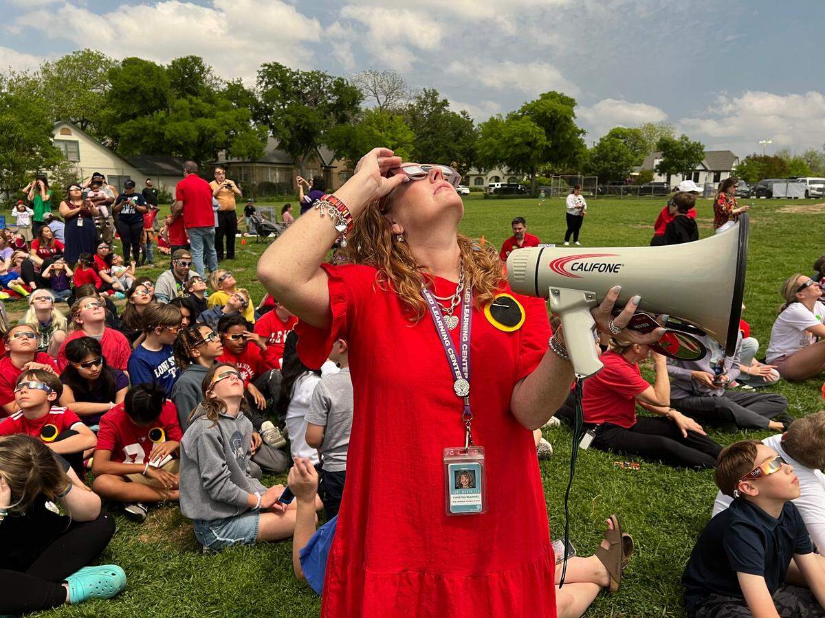 A Fort Worth ISD teacher is standing among a crowd of elementary students who are gazing at the sky wearing protective eyeglasses during the solar eclipse. She is standing while holding her glasses in place, and students are sitting down on a grassy field.